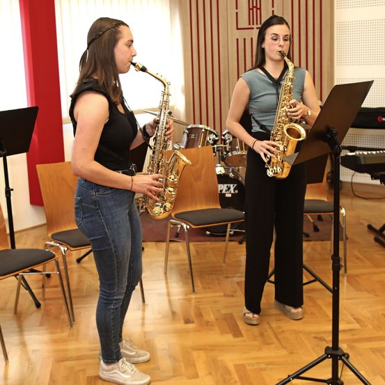 Zwei Frauen spielen Saxophon in einem Studio mit Holzboden. Eine steht mit einem Notenständer, die andere lehnt an einer roten Wand.