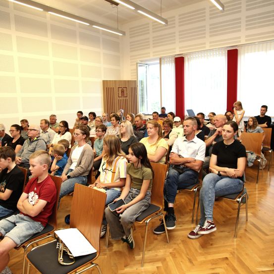 Eine große Gruppe von Menschen unterschiedlichen Alters sitzt in Reihen von Stühlen in einem Konferenzraum. Ein Trompete liegt auf einem Stuhl vor ihnen.