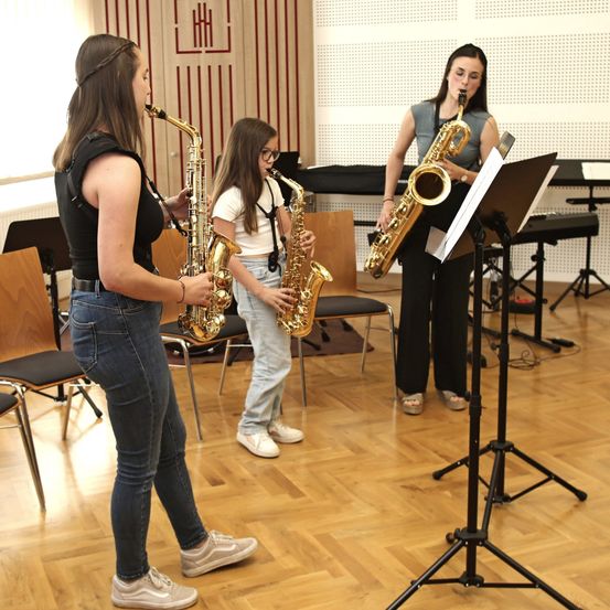 Drei Frauen in einem Musikraum, jede hält ein Saxophon und eine liest Noten. Zwei stehen und eine sitzt auf einem Stuhl. Sie sind lässig gekleidet.