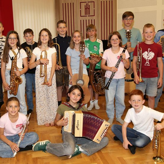 Eine Gruppe von Kindern und Jugendlichen posiert mit verschiedenen Musikinstrumenten. Sie sitzen und stehen in einem Halbkreis. Zu den Instrumenten gehören Trompeten, Klarinetten, Saxophone und Gitarren.