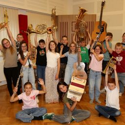 Eine Gruppe junger Musiker posiert mit verschiedenen Instrumenten, darunter Trompeten, Saxophone und Gitarren, in einem Musikstudio.