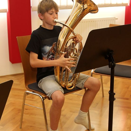 Ein junger Junge sitzt auf einem Stuhl und spielt eine Messingtuba, während er das Notenblatt auf einem Ständer vor sich betrachtet.