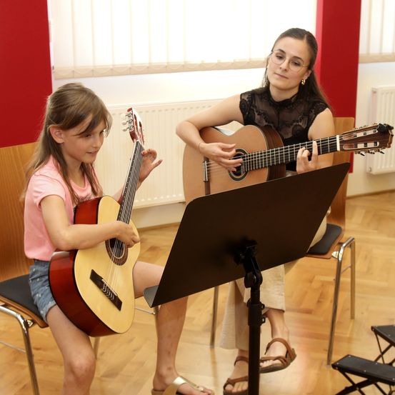 Zwei Mädchen spielen Gitarre in einem Studio mit Holzboden. Eine sitzt, und die andere steht hinter einem Notenständer.