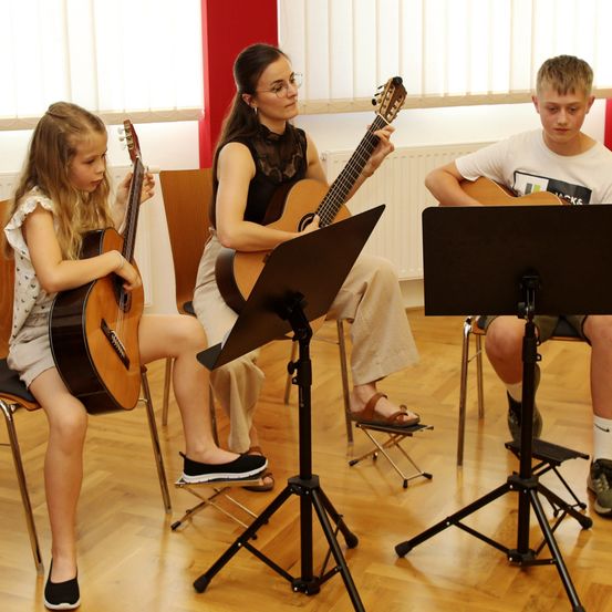Drei Kinder spielen Gitarre in einem Raum mit Holzboden und Heizkörpern. Das Mädchen und die Frau tragen Sandalen, der Junge trägt Turnschuhe.