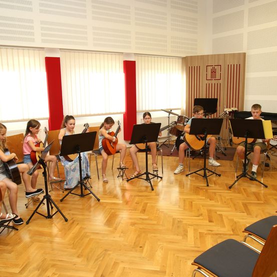 Eine Gruppe von Kindern übt Gitarre in einem Raum mit Holzböden, roten Säulen und Fenstern. Sie sitzen auf Stühlen und spielen Gitarren vor Musikständern.