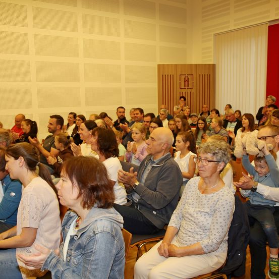 Eine große Gruppe von Menschen ist in einem Raum versammelt, klatscht und lächelt. Einige sitzen auf Stühlen, andere stehen. Der Raum hat weiße Wände, eine rote Fahne und eine Holztür.