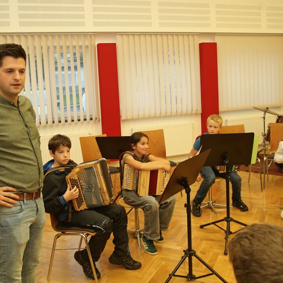 Vier junge Kinder spielen Akkordeons in einem Klassenzimmer. Ein Mann steht vor ihnen. Hinter ihnen sind Musikinstrumente aufgestellt.