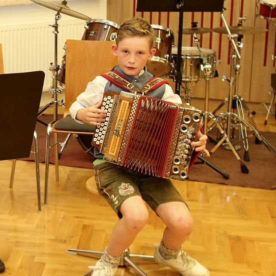 Ein junger Junge spielt ein Akkordeon in einem Musikstudio, umgeben von Trommeln und Becken.
