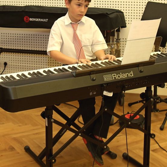 Ein Junge in einem weißen Hemd und einer Krawatte spielt ein Roland-Keyboard und hält ein Musikblatt. Er sitzt auf einem Hocker mit einem Holzboden und einem Notenständer hinter ihm.