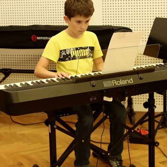 Ein junger Junge sitzt an einem schwarzen Klavier und trägt ein gelbes T-Shirt mit einem schwarzen und weißen Grafikdesign. Er liest Notenblätter, die auf dem Klavier liegen. Hinter ihm ist ein schwarzer Stoffvorhang zu sehen.