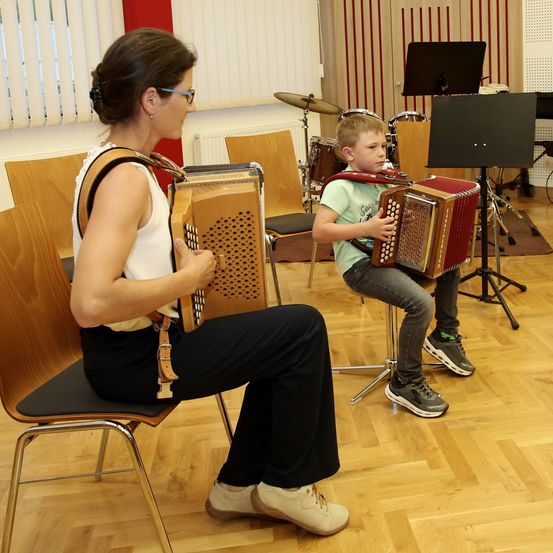 Eine Frau und ein junger Junge spielen Akkordeon in einem Raum mit Holzboden. Dahinter sind Stühle, Trommeln und ein Notenständer.