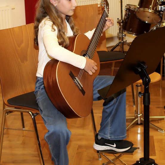Ein Mädchen sitzt auf einem Stuhl in einem Musikzimmer, spielt eine Gitarre und liest Noten auf einem Ständer. Hinter ihr stehen ein Schlagzeug und ein Holzstuhl.