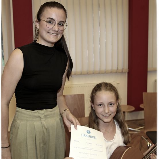 Eine Frau und ein junges Mädchen halten ein Zertifikat. Die Frau trägt eine Brille und ein schwarzes ärmelloses Oberteil. Das Mädchen hält eine Gitarre. Sie stehen vor einer weißen Wand mit Jalousien und einem Holzstuhl.