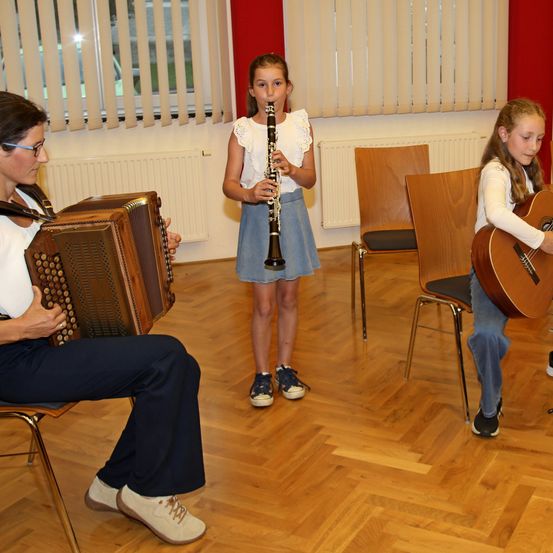 Drei Personen, darunter zwei junge Mädchen und ein Erwachsener, spielen Musikinstrumente in einem Raum mit Holzböden und Stühlen. Ein Mädchen spielt Klarinette, das andere Gitarre. Ein Erwachsener spielt Akkordeon.