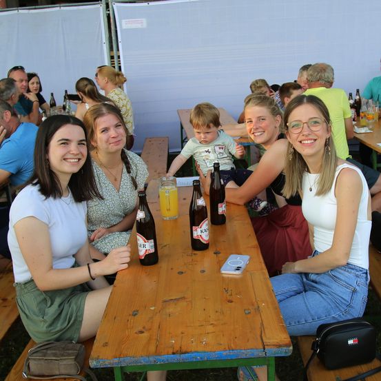 Eine Gruppe von Frauen sitzt an einem Tisch in einem Biergarten im Freien, mit Bierflaschen, einem Glas Orangensaft und einem Handy auf dem Tisch. Sie lächeln und scheinen sich zu amüsieren.