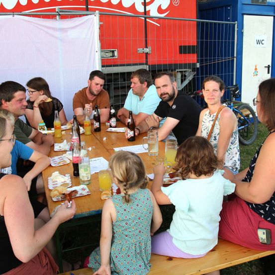Eine Gruppe von Erwachsenen und Kindern sitzt im Freien um einen Tisch herum und genießt Essen und Getränke. Der Tisch ist mit Flaschen, Gläsern und Tellern gefüllt. Ein Fahrrad ist in der Nähe geparkt. Dahinter steht ein roter LKW mit einem weißen Banner.