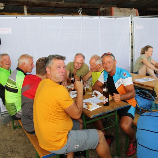 Eine Gruppe älterer Männer in Radsporttrikots und -shorts sitzt um einen Tisch mit Flaschen und Essen, wahrscheinlich während einer Pause bei einer Radsportveranstaltung.