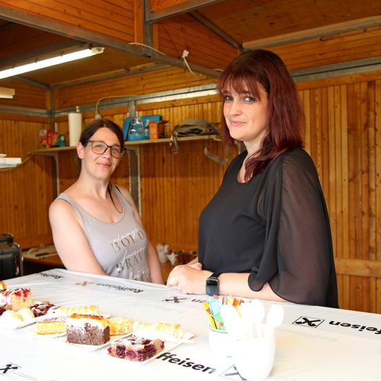 Zwei Frauen stehen hinter einem Tisch mit verschiedenen Desserts. Eine Frau trägt eine Brille und ein ärmelloses Shirt, die andere eine schwarze Bluse. Hinter ihnen stehen Holzregale mit verschiedenen Gegenständen.