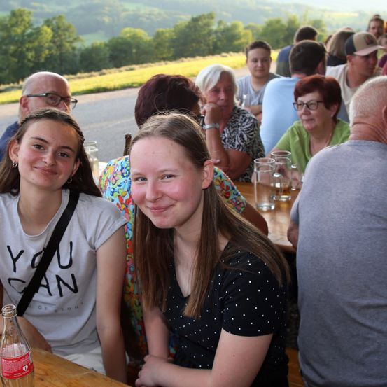 Eine Gruppe von Menschen sitzt an einem Tisch im Freien, zwei junge Frauen lächeln. Bäume und eine Straße sind im Hintergrund zu sehen.