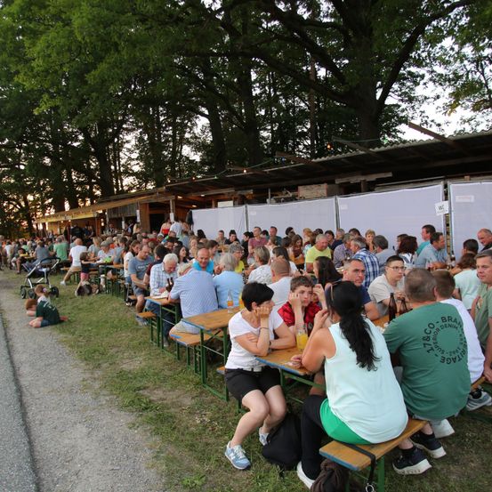 Eine Menschenmenge sitzt an Tischen in einem Außenbereich, umgeben von Bäumen und Gras. Einige essen oder trinken. Ein Kinderwagen steht in der Nähe.
