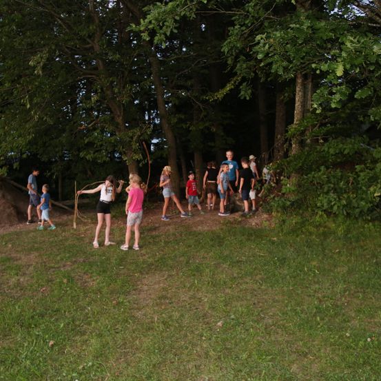 Eine Gruppe von Kindern im Wald, einige halten Stöcke, andere beobachten. Der Grasbereich ist von Bäumen und Pflanzen umgeben.