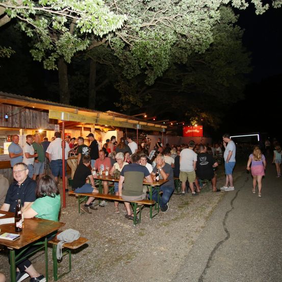 Eine Nachtveranstaltung in einem Außenbereich einer Bar mit vielen sitzenden und stehenden Personen, die Getränke genießen und sich unterhalten, unter Bäumen.