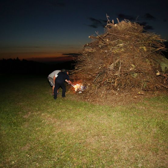 Zwei Personen zünden bei Nacht ein Feuer in der Nähe eines großen Haufens Zweige und Äste an.