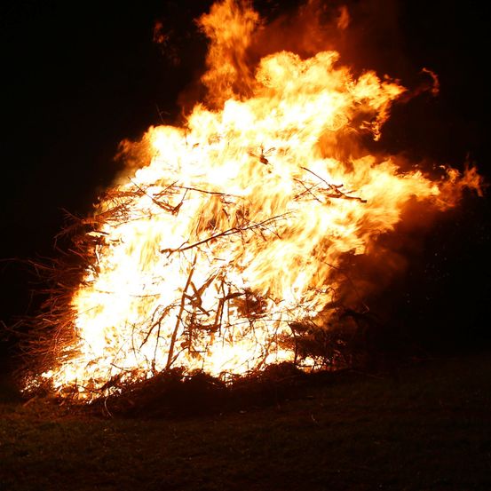 Ein großes Lagerfeuer brennt in der dunklen Nacht hell und wirft flackernde Schatten auf die Umgebung.