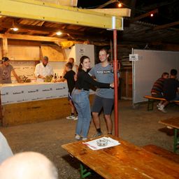 Ein Mann und eine Frau tanzen nachts in einer Bar. Hinter ihnen steht ein Mann in einer Kochjacke am Tresen. Leute sitzen auf Bänken.
