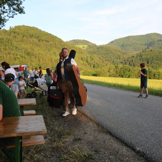 Ein Mann, der einen Gitarrenkoffer trägt, geht einen Feldweg entlang, mit einer Menschenmenge im Hintergrund und Bergen in der Ferne.