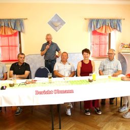 Eine Gruppe von Menschen sitzt um einen langen Tisch, an dem ein Mann steht und in ein Mikrofon spricht. Der Tisch ist mit einer weißen Tischdecke bedeckt und hat Gläser und ein Buch darauf. Der Raum hat gelbe Wände und rote Vorhänge.