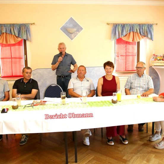 Eine Gruppe von Menschen sitzt um einen langen Tisch, an dem ein Mann steht und in ein Mikrofon spricht. Der Tisch ist mit einer weißen Tischdecke bedeckt und hat Gläser und ein Buch darauf. Der Raum hat gelbe Wände und rote Vorhänge.