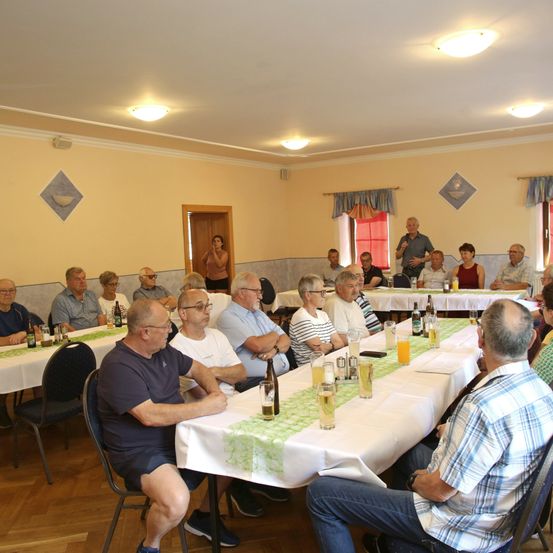 Eine Gruppe von Menschen trifft sich in einem Raum für eine Versammlung, sitzt um Tische mit Getränken und Essen. Einige tragen Brillen und Hüte. Eine Frau steht in der Nähe der Tür.