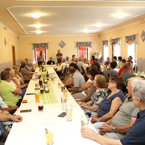 Eine Gruppe von Menschen sitzt an einem langen Tisch in einem Speisesaal, mit mehreren Gläsern Bier und Flaschen. Ein Mann steht vorne, möglicherweise hält er eine Rede.
