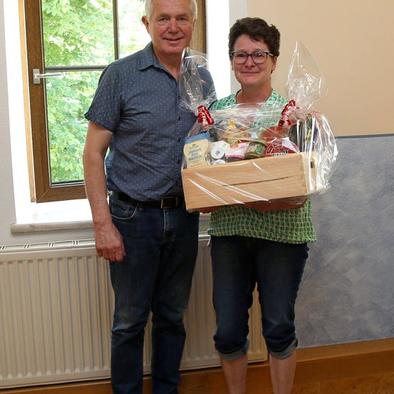 Ein Mann und eine Frau stehen nebeneinander und halten eine Holzkiste mit verschiedenen Lebensmitteln. Der Mann trägt ein blaues Hemd und Jeans, die Frau trägt ein grünes Hemd und eine Brille. Sie stehen vor einem Fenster mit Bäumen draußen.