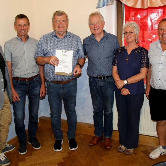 Fünf Personen stehen lächelnd zusammen in einem Raum. Der Mann in der Mitte hält ein Zertifikat. Hinter ihnen ist ein roter Vorhang und eine weiße Wand zu sehen.