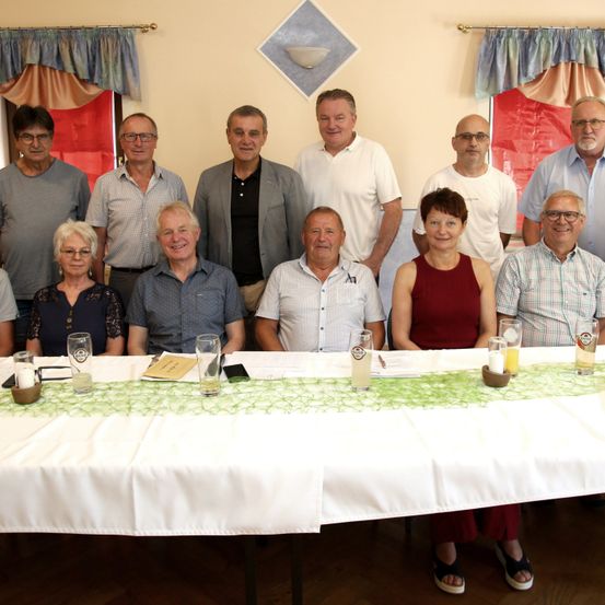 Eine Gruppe von Menschen, sowohl Männer als auch Frauen, sitzt um einen Tisch in einem Raum. Sie lächeln und scheinen für ein Foto zu posieren. Der Tisch ist mit einem weißen Tuch bedeckt und hat Gläser, eine Kerze und ein Menü.