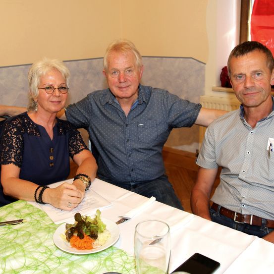 Drei Personen sitzen an einem Tisch und lächeln für ein Foto. Eine Frau mit Brille und blauer Bluse, ein Mann in einem blauen Hemd mit einem Stift in der Tasche und ein anderer Mann in einem grauen Hemd. Ein Teller mit Salat und ein Glas Wasser stehen auf dem Tisch.