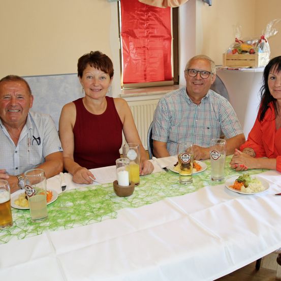 Vier Personen sitzen an einem Tisch mit Tellern mit Essen und Getränken und lächeln für ein Foto. Der Tisch ist mit einer grünen Tischdecke und einer weißen Tischplatte gedeckt. Hinter ihnen befindet sich ein Fenster mit einem roten Vorhang.