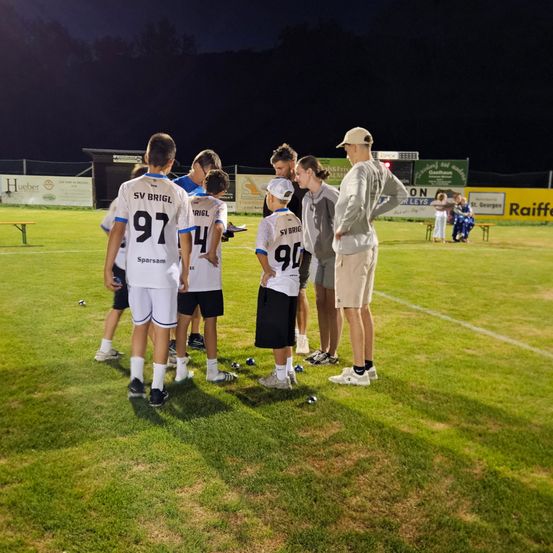 Eine Gruppe von Fußballspielern in weißen Trikots steht nachts auf einem Feld zusammengedrängt, wobei ein Trainer in einer beigefarbenen Kappe und Turnschuhen die Diskussion leitet.