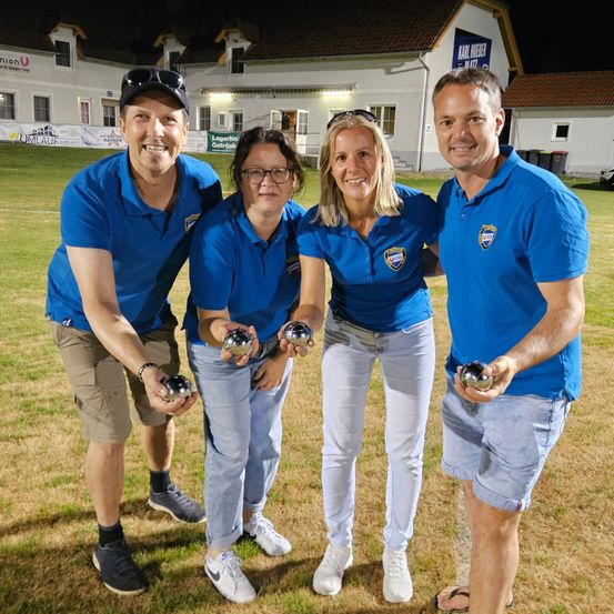 Vier Personen in blauen Hemden posieren für ein Foto auf einem Grasfeld bei Nacht und halten silberne Kugeln. Hinter ihnen steht ein Gebäude mit einem Schild, das 'Karl Weber' besagt.