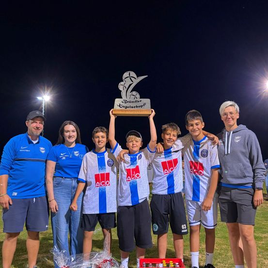 Eine Gruppe junger Jungen und ihre Trainer feiern mit einem Pokal auf einem Fußballfeld bei Nacht.