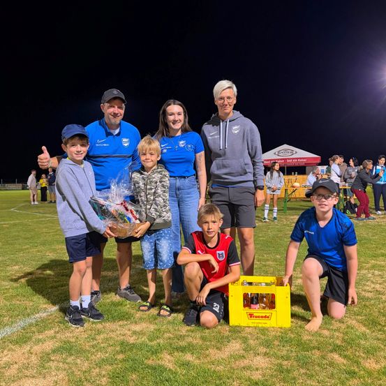 Eine Gruppe von Menschen, einschließlich Erwachsener und Kindern, posiert für ein Foto auf einem Grasfeld bei Nacht. Sie halten eine gelbe Box und eine Plastiktüte mit Gegenständen darin. Hinter ihnen stehen Zelte und Menschen. Der Himmel ist dunkel.
