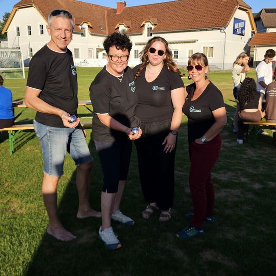Vier Personen in passenden schwarzen T-Shirts stehen auf einem Rasenfeld, lächeln und halten Metallkugeln. Hinter ihnen ist ein Gebäude mit einem Schild zu sehen, das 'Karl Huber Platz' besagt.