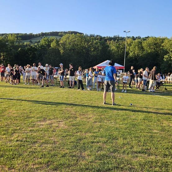 Eine Gruppe von Menschen versammelt sich auf einem Rasenfeld, möglicherweise für eine Sportveranstaltung. Einige stehen, andere sitzen auf Stühlen. Zelte und Tische sind im Bereich aufgestellt. In der Ferne gibt es Bäume und eine Bergkette.