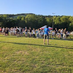 Eine Gruppe von Menschen versammelt sich auf einem Rasenfeld, möglicherweise für eine Sportveranstaltung. Einige stehen, andere sitzen auf Stühlen. Zelte und Tische sind im Bereich aufgestellt. In der Ferne gibt es Bäume und eine Bergkette.