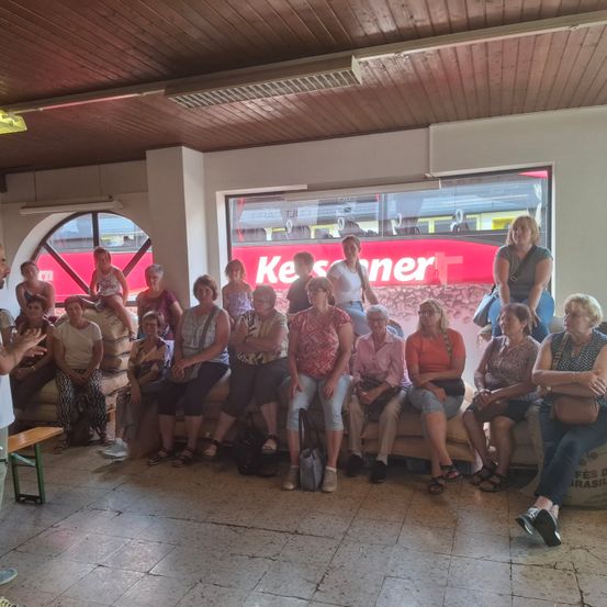 Eine Gruppe von Frauen und einem Mann sitzt auf einer Bank in einem Raum mit einer Holzdecke und einem großen Fenster, das ein rotes Schild zeigt.