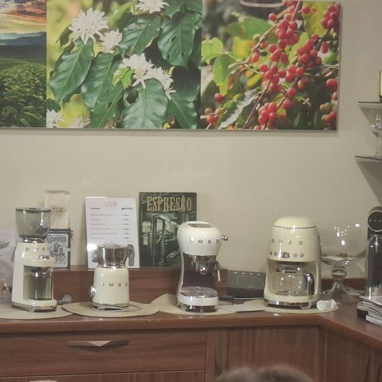 Ein Café-Schalter zeigt verschiedene Kaffeemaschinen und ein Buch mit dem Titel Espresso. Darüber befinden sich drei Poster von Kaffeepflanzen und -bohnen an der Wand.