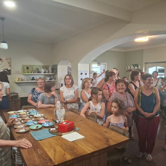 Eine Gruppe von Frauen und Kindern versammelt sich um einen Tisch mit verschiedenen Tellern und Utensilien. Sie scheinen sich auf eine Veranstaltung vorzubereiten.