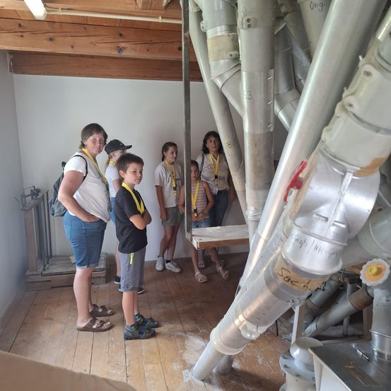 Eine Gruppe von Menschen, einschließlich Erwachsener und Kinder, steht in einem Raum mit Holzböden und großen Metallrohren. Sie tragen gelbe Lanyards und scheinen auf einer Tour zu sein.